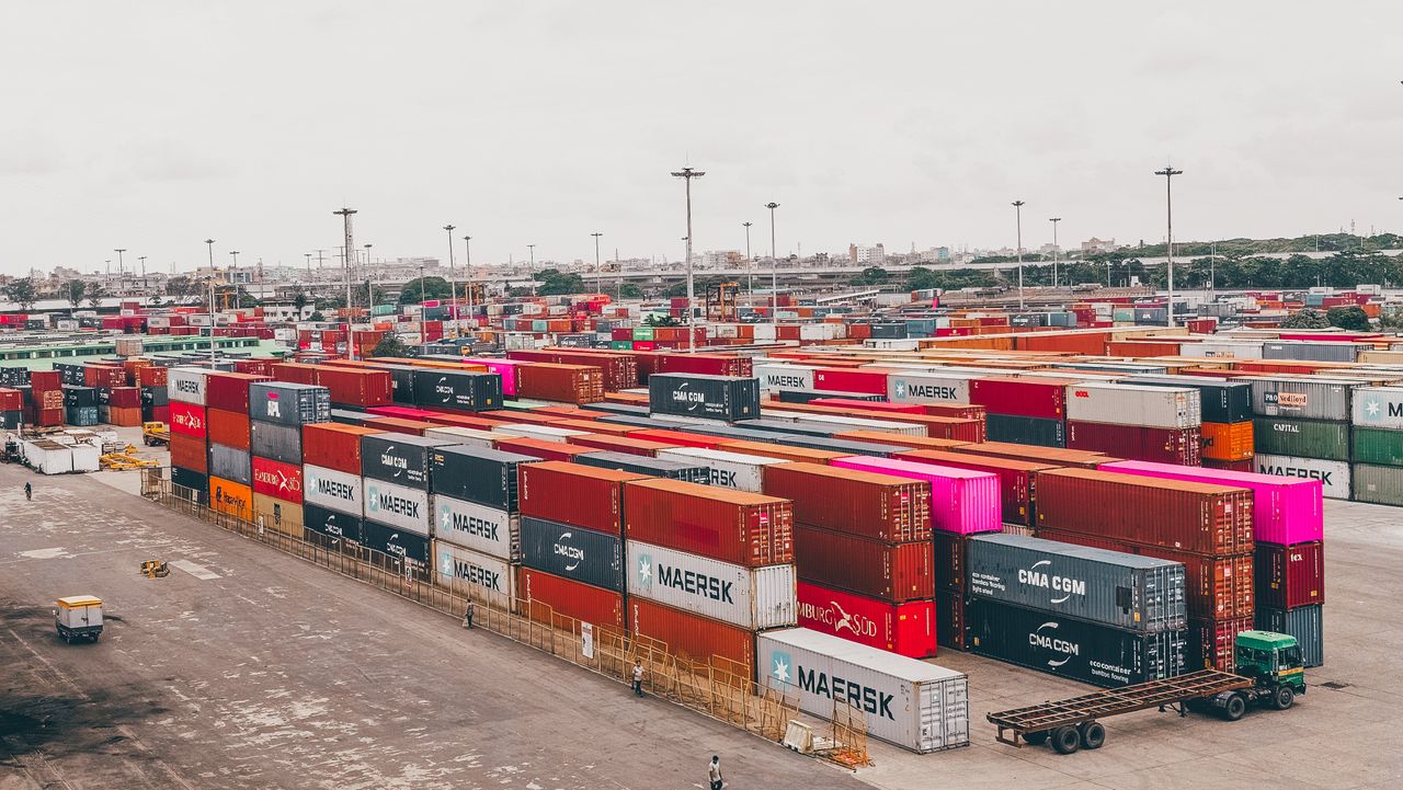 hero-img-01 Colorful shipping containers stacked at a bustling port showcasing global trade.