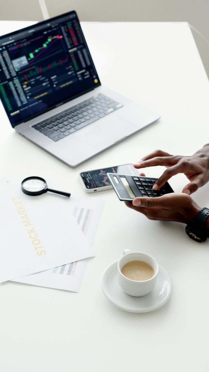 gallery-1 A businessman using a laptop and smartphone to analyze stock market trends indoors.